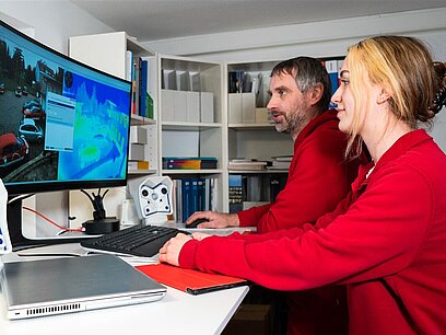 Eine junge Frau und ein älterer Mann in Arbeitskleidung bearbeiten am PC eine Aufgabe. 